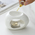 White ceramic oil burner with a lit candle and a hand dropping oil into it.