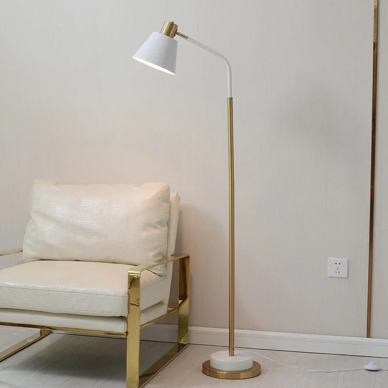 White Nordic Floor Lamp — White Nordic-style metal floor lamp with gold-tone stem, angled shade, and round base, shown beside a living room chair.