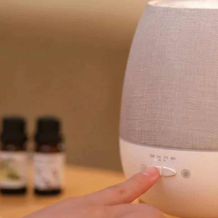 White diffuser with gray top on a wooden surface, hand pressing a button, essential oils in the background.