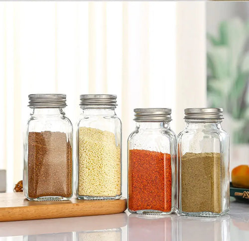Four glass jars filled with colourful spices including chili powder and cumin, displayed on a wooden board.