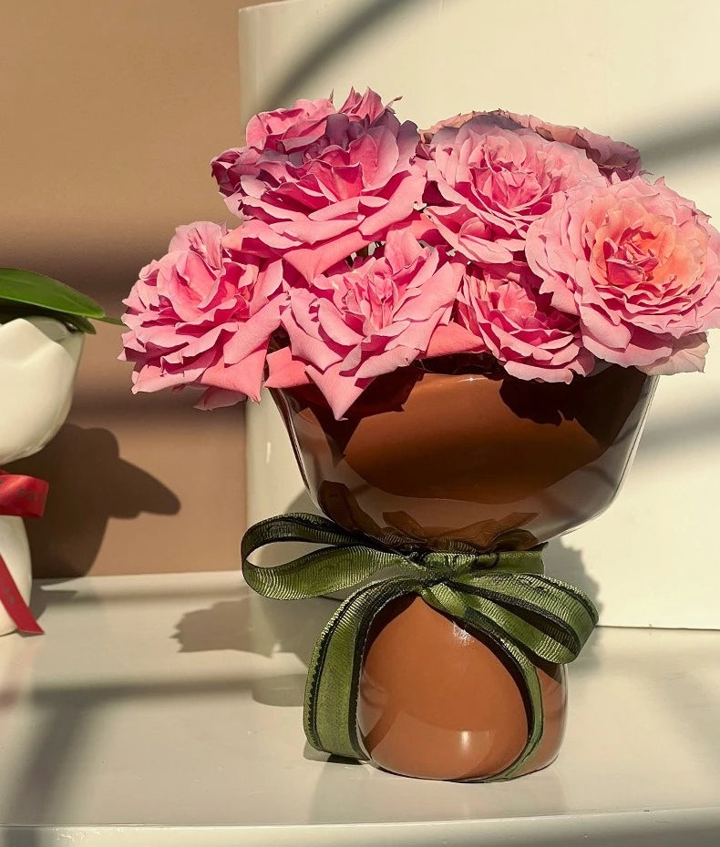 Bouquet of pink flowers in a brown vase with a green ribbon on a white surface.