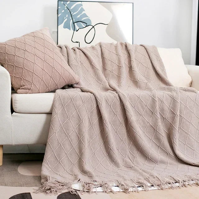 Taupe knit throw with a diamond texture and tassels draped over a sofa beside a matching cushion
