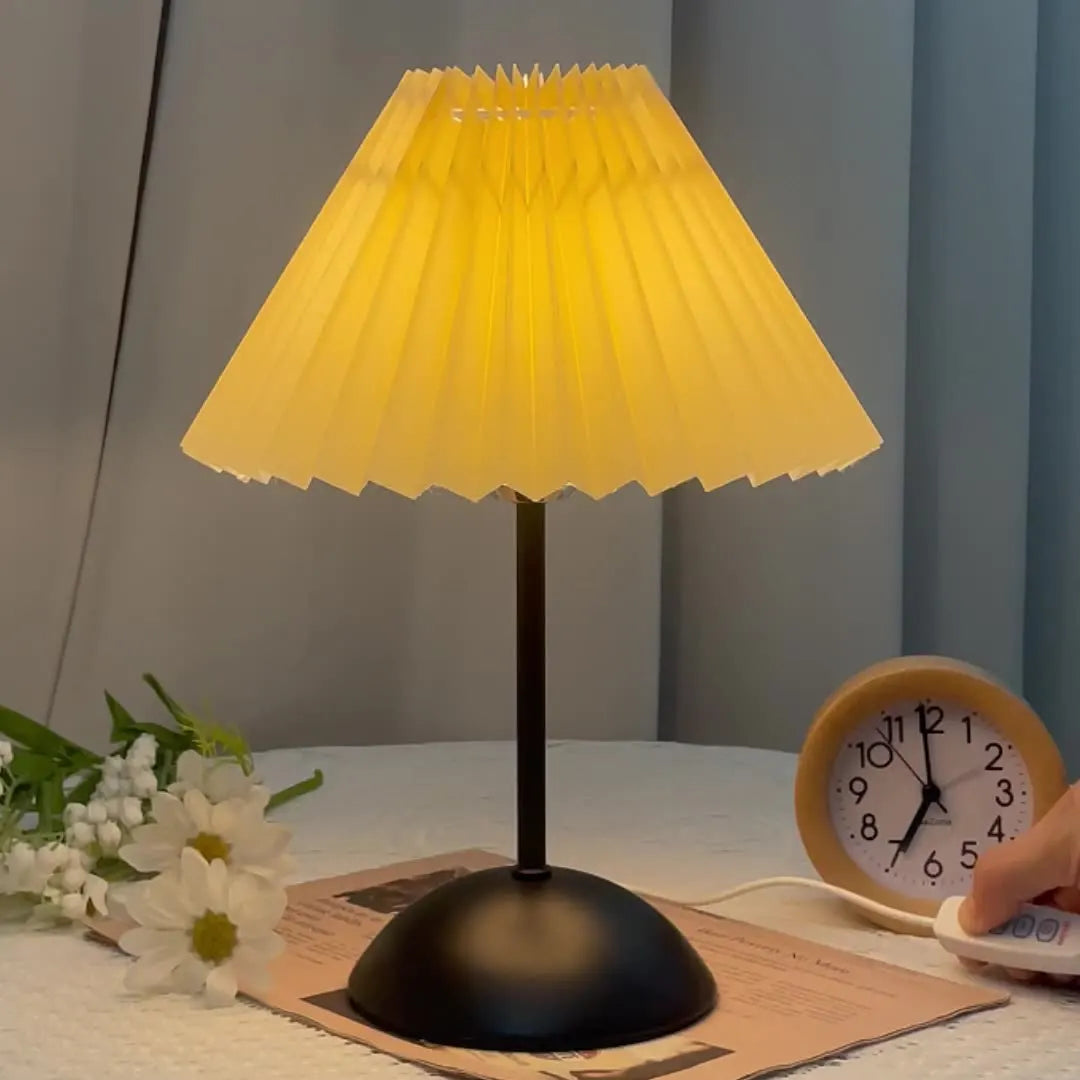 Yellow pleated lampshade on a black base with a clock and flowers on a table.