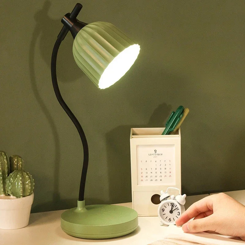 Green desk lamp on a table with a hand holding a book, cactus plant, and calendar in the background.