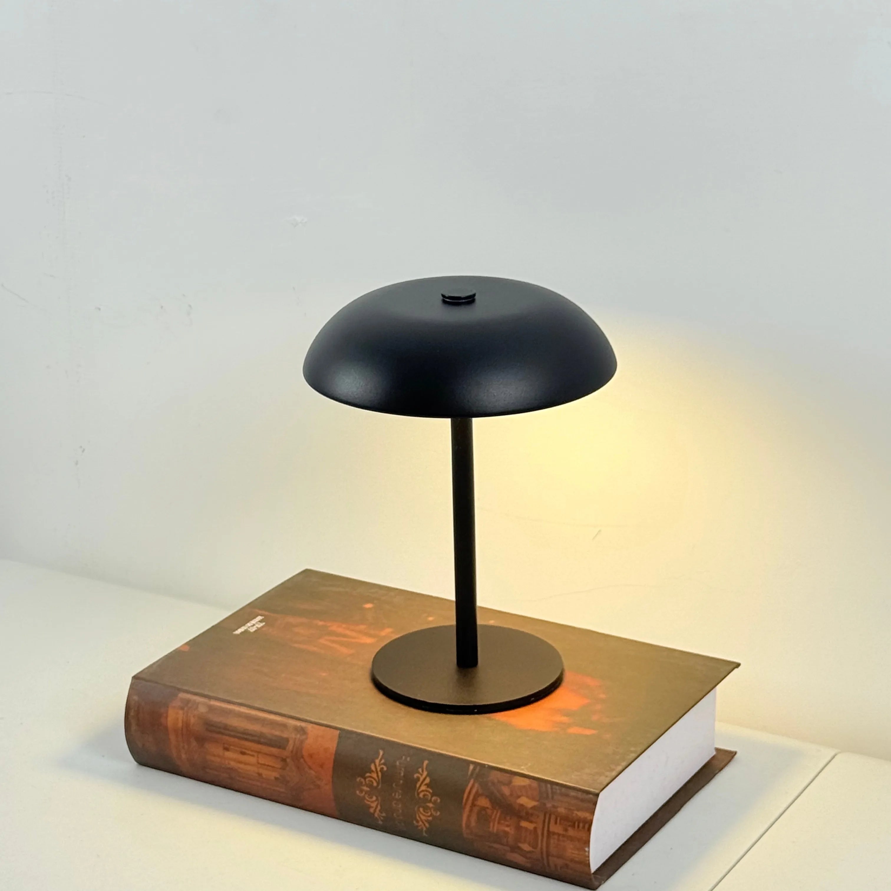 Black mushroom lamp with a round shade on top of a book against a light gray background