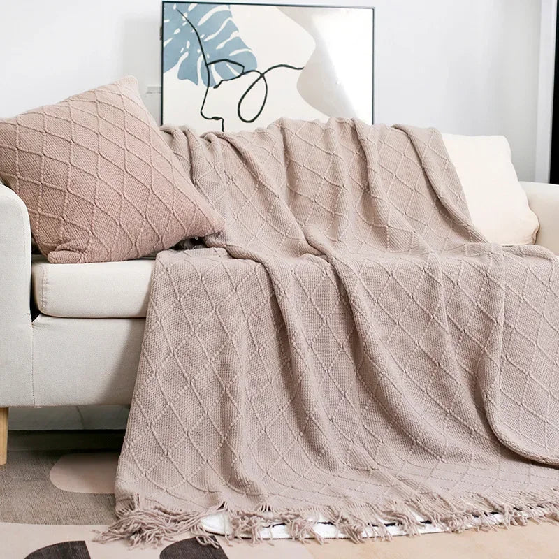Taupe knit throw with a diamond texture and tassels draped over a sofa beside a matching cushion