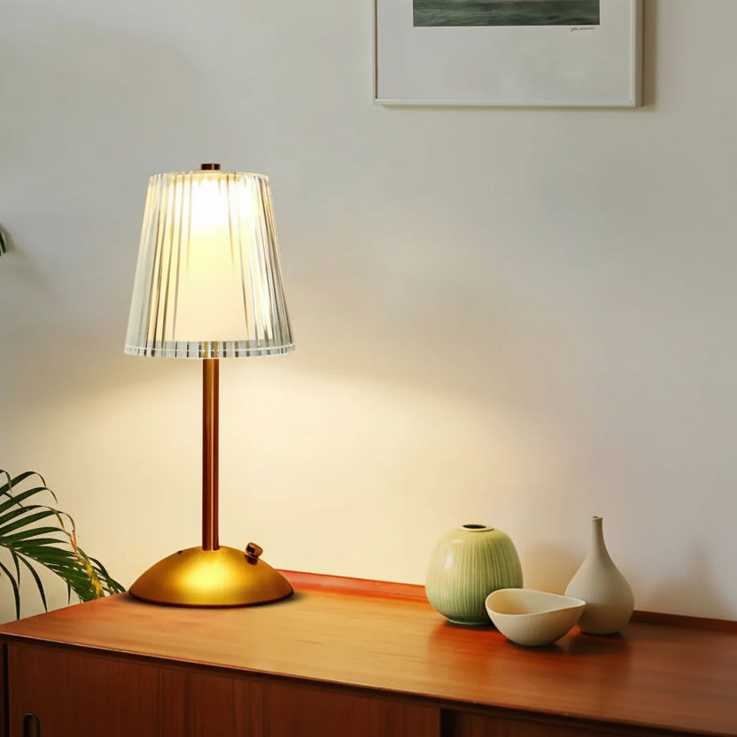 Gold table lamp with white shade on a wooden surface, surrounded by decorative items and a plant.