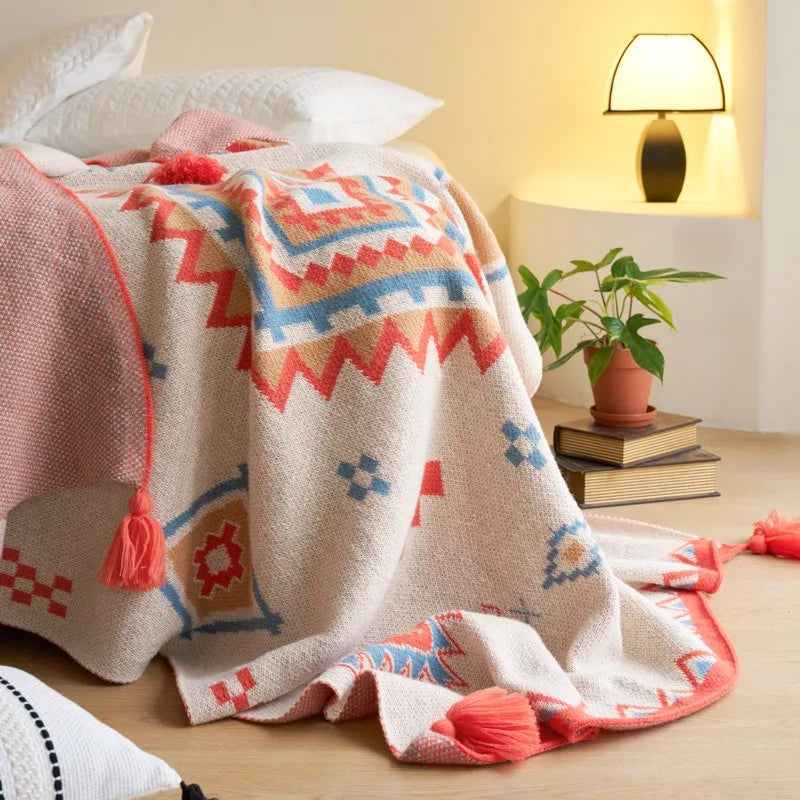 A woven throw blanket with a Southwestern geometric pattern in terracotta, blue, and cream is draped over a neatly made bed in a bright, sunlit bedroom.
