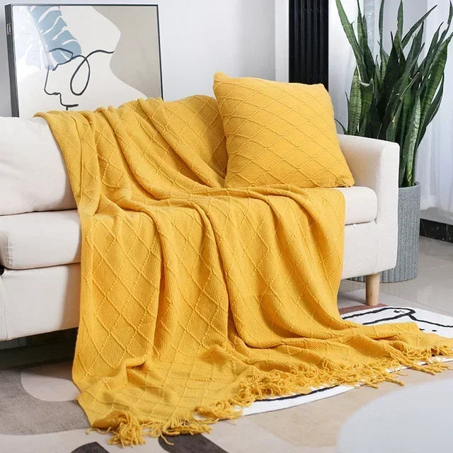 Mustard yellow diamond-textured throw with fringe laid across a cream sofa, with a matching cushion and green plant nearby