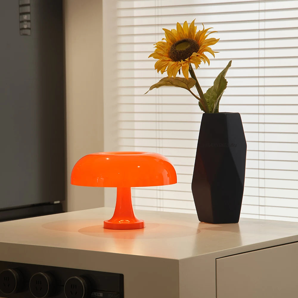 Orange lamp and black vase with sunflower on a surface near a window with blinds.