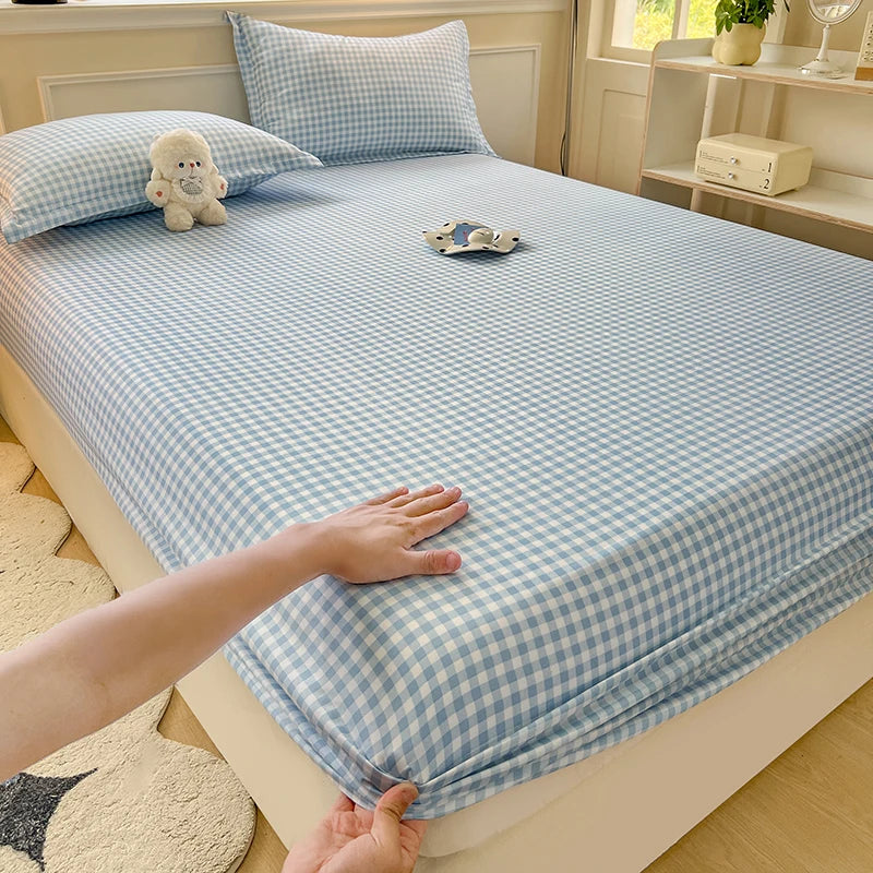 A bed made with a light blue and white gingham check fitted sheet and pillowcase. A small white plush bear is resting on the pillow in a brightly lit, aesthetic room.