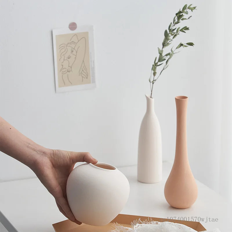 Hand holding a white ceramic vase with two other vases on a light surface.