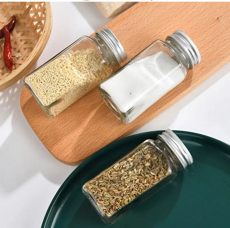 Three glass spice jars with silver lids on a wooden board and green plate.