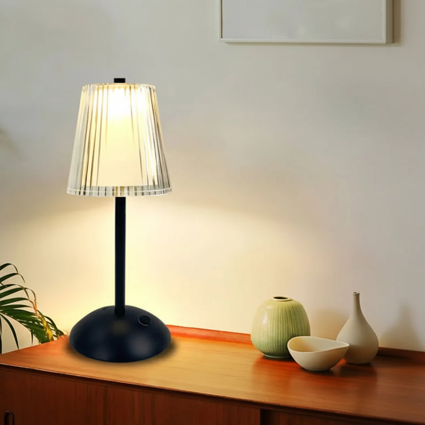 Table lamp with white shade on a wooden surface, surrounded by decorative items and a plant.