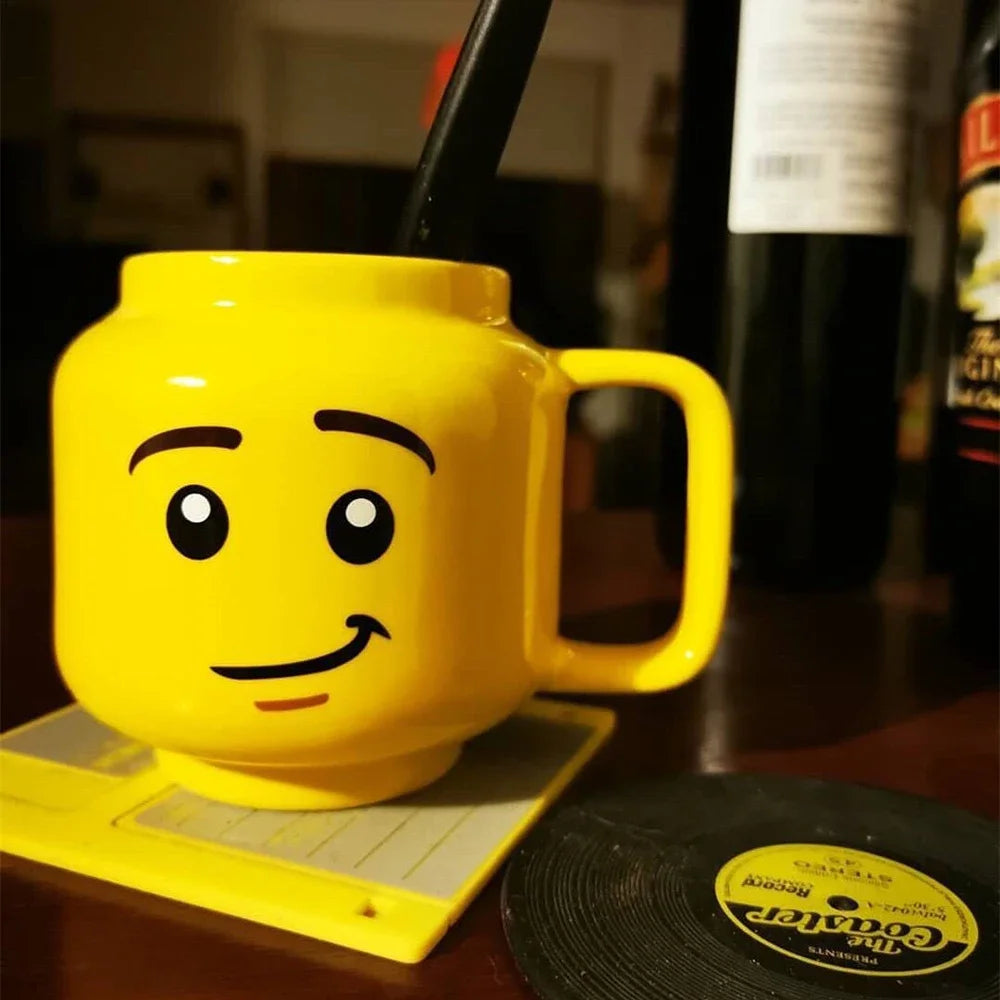 Yellow mug shaped like a lego figure head on a table with bottles in the background