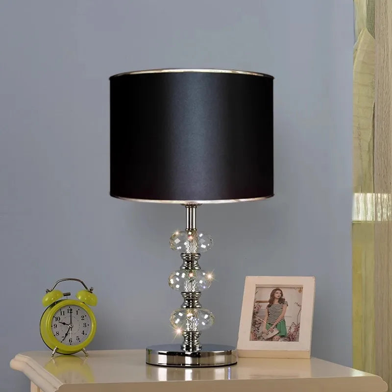 table lamp with a solid black cylindrical shade and a triple-stacked crystal base on a nightstand.