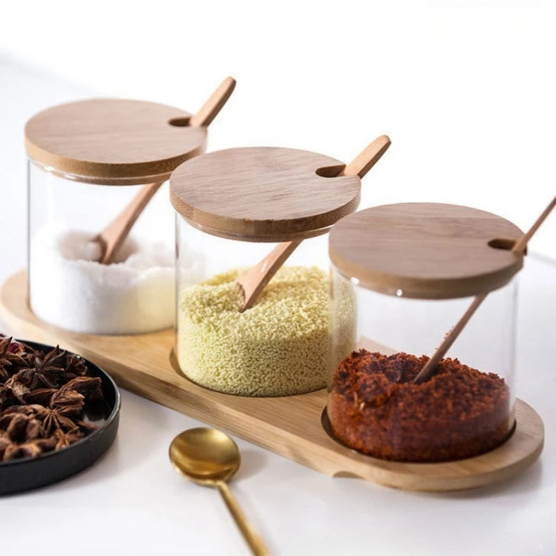 Three glass jars with wooden lids and spoons on a wooden tray, containing different spices.