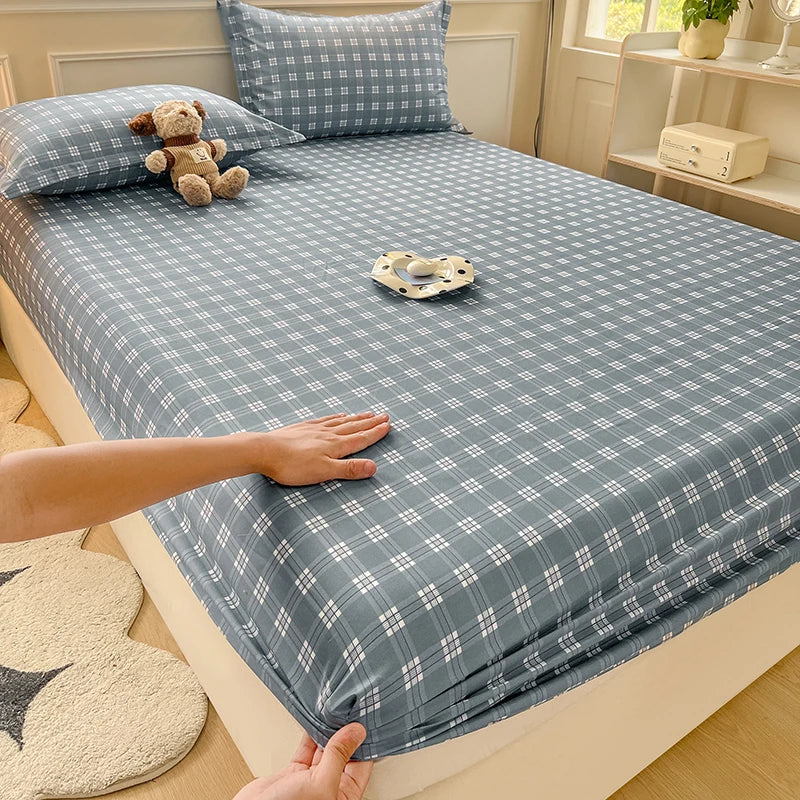 A bed styled with a dusty blue and grey plaid fitted sheet. Sunlight filters into the room, highlighting the bedding's clean, aesthetic lines.