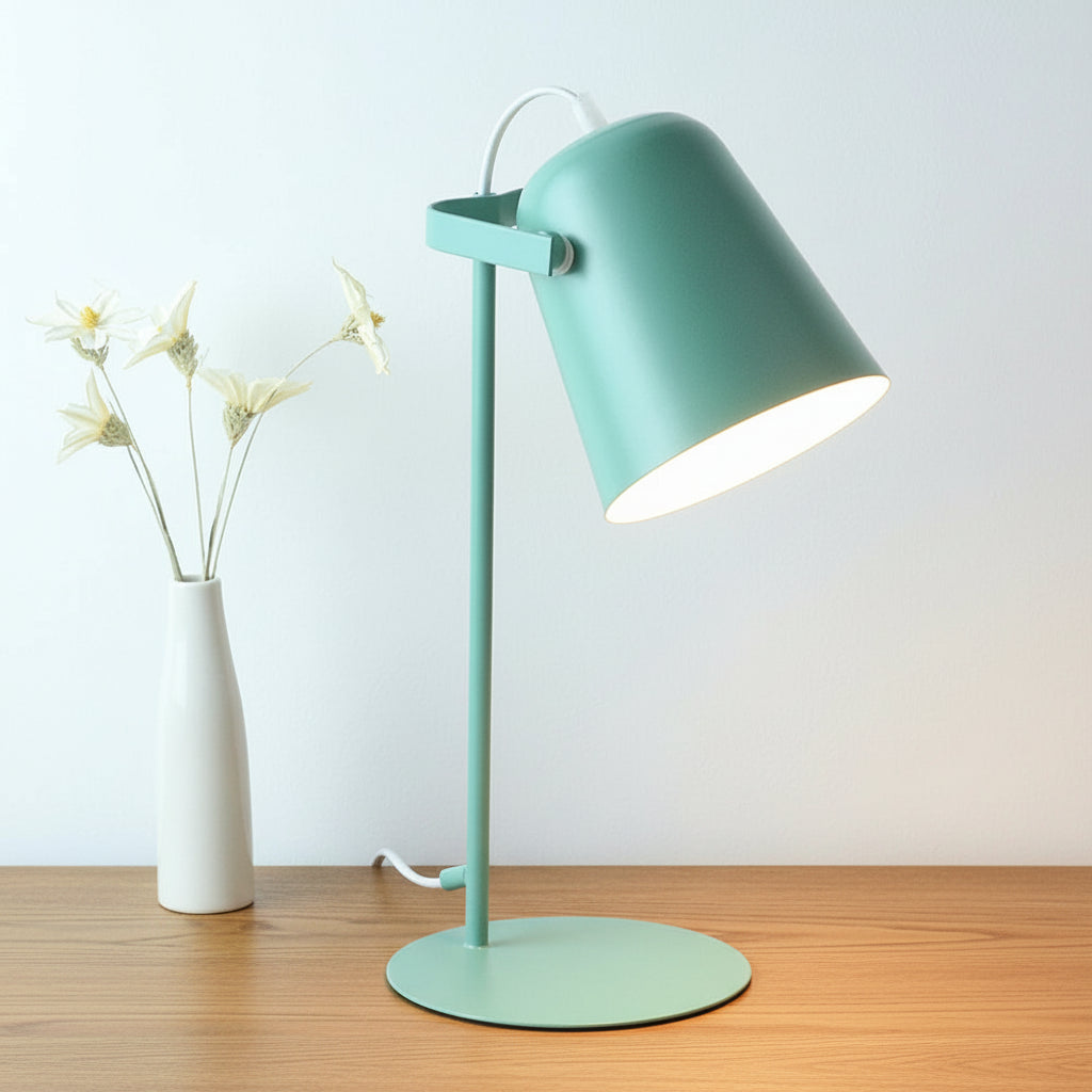 Mint green desk lamp on a white surface with a white vase containing dried flowers in the background.