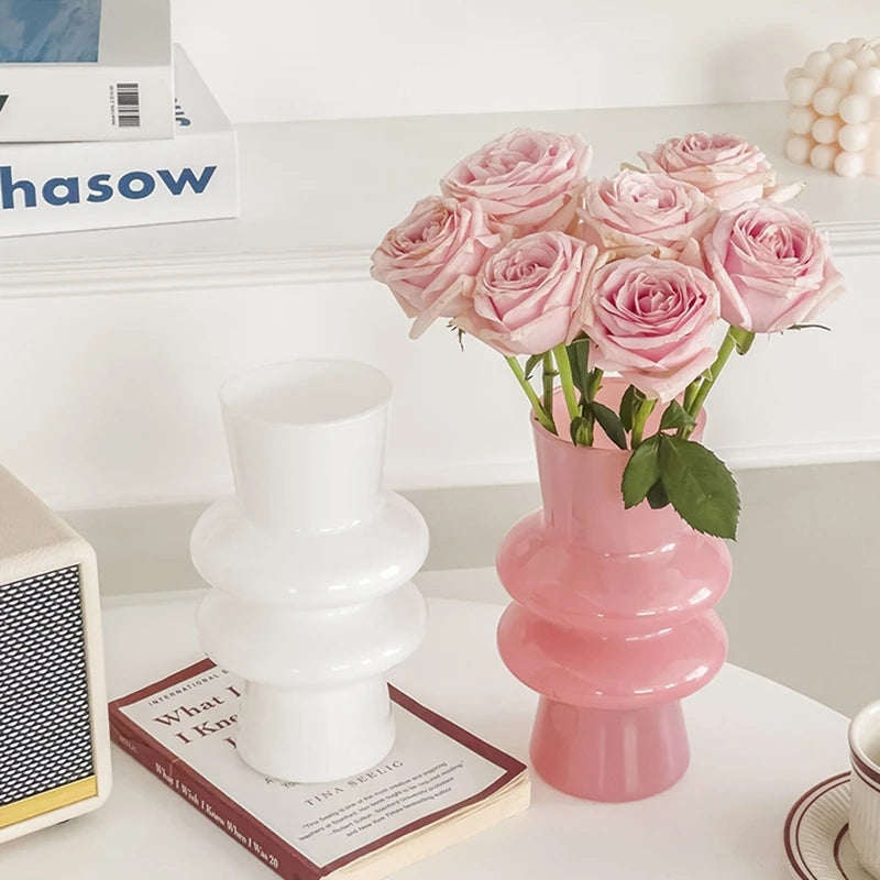 Pink and white vase with pink roses on a white surface, with books and a lamp.