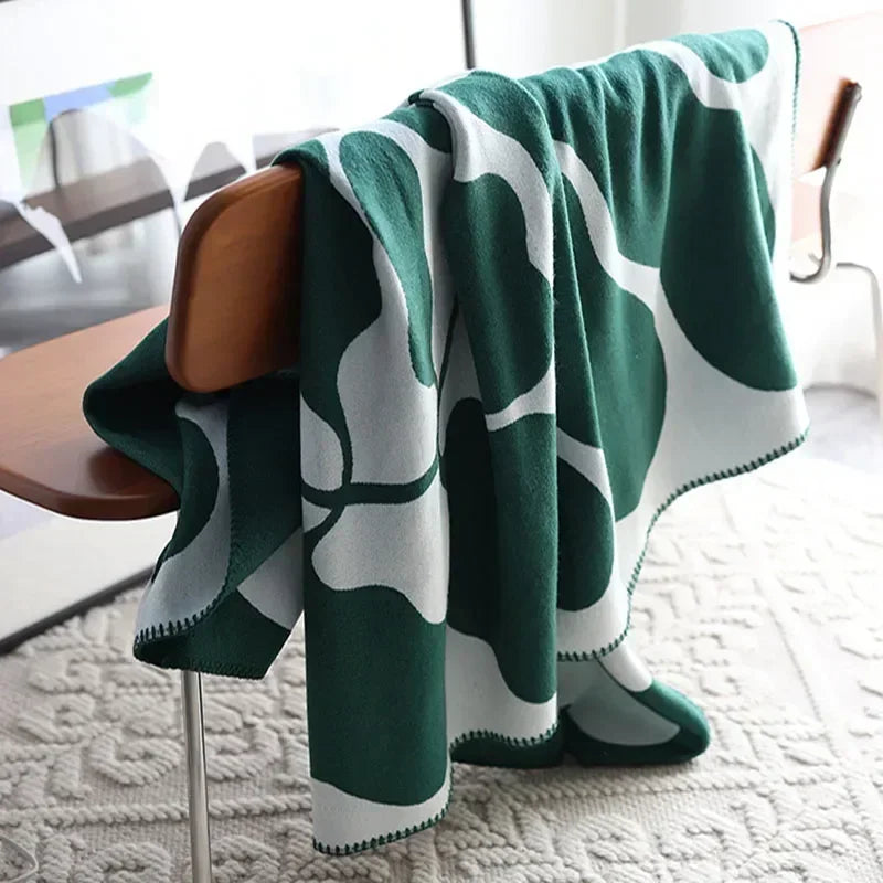 Forest green and white abstract jacquard blanket with blanket stitch edge draped over a wooden chair on a textured cream rug