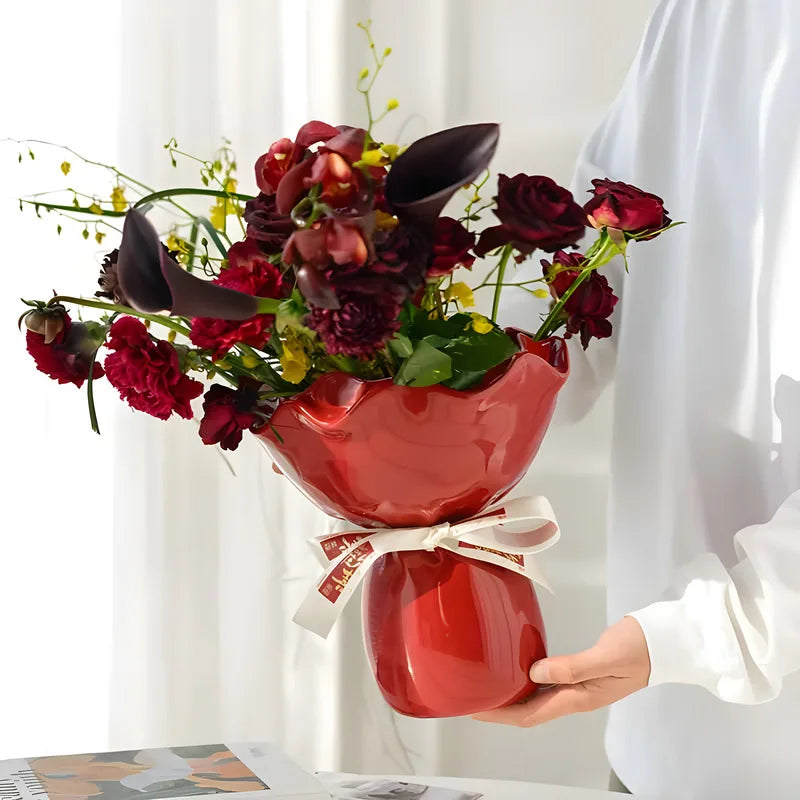 Person holding a bouquet of red and dark purple flowers in a red vase against a white curtain background.