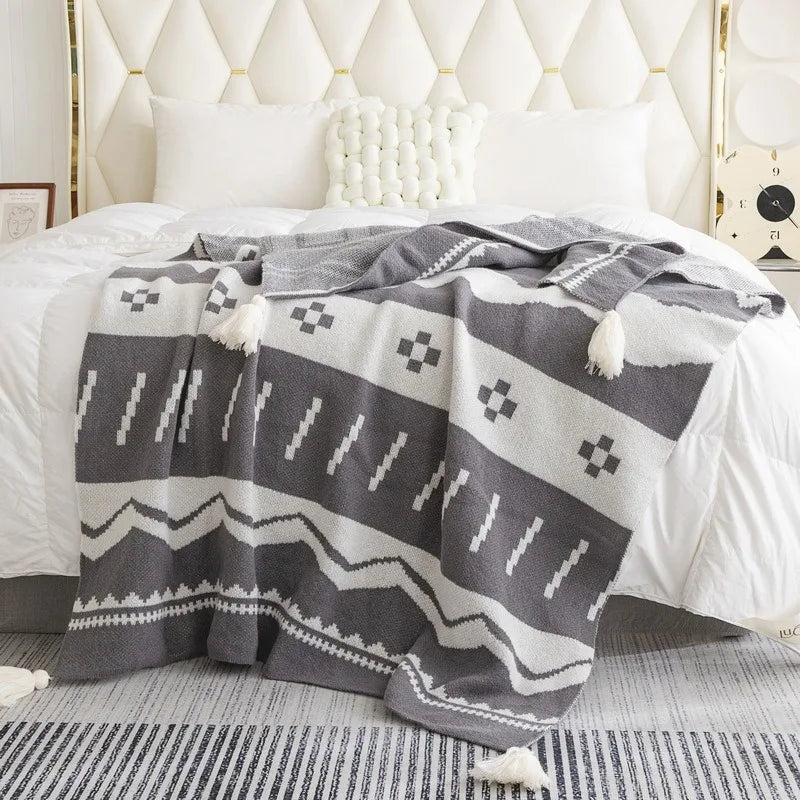 A woven throw blanket with a dark gray and cream geometric pattern and tassels, draped over a bed in a bright, modern bedroom.