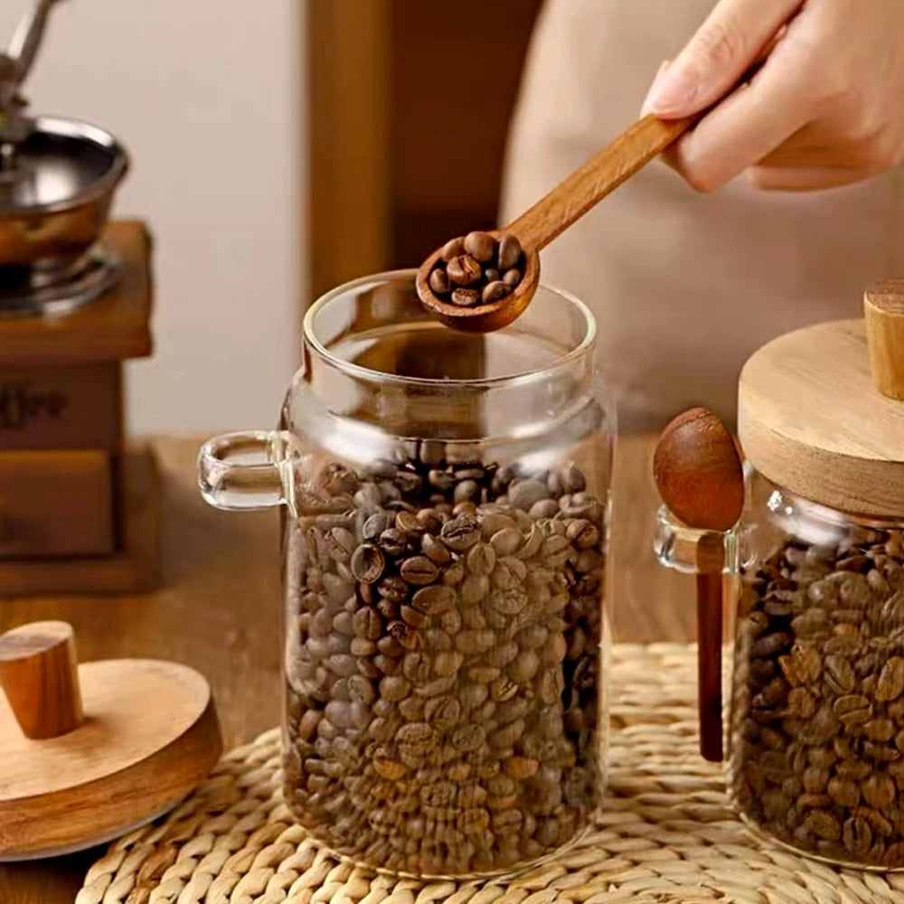 Glass jar filled with coffee beans on a woven mat, with a hand holding a wooden scoop.