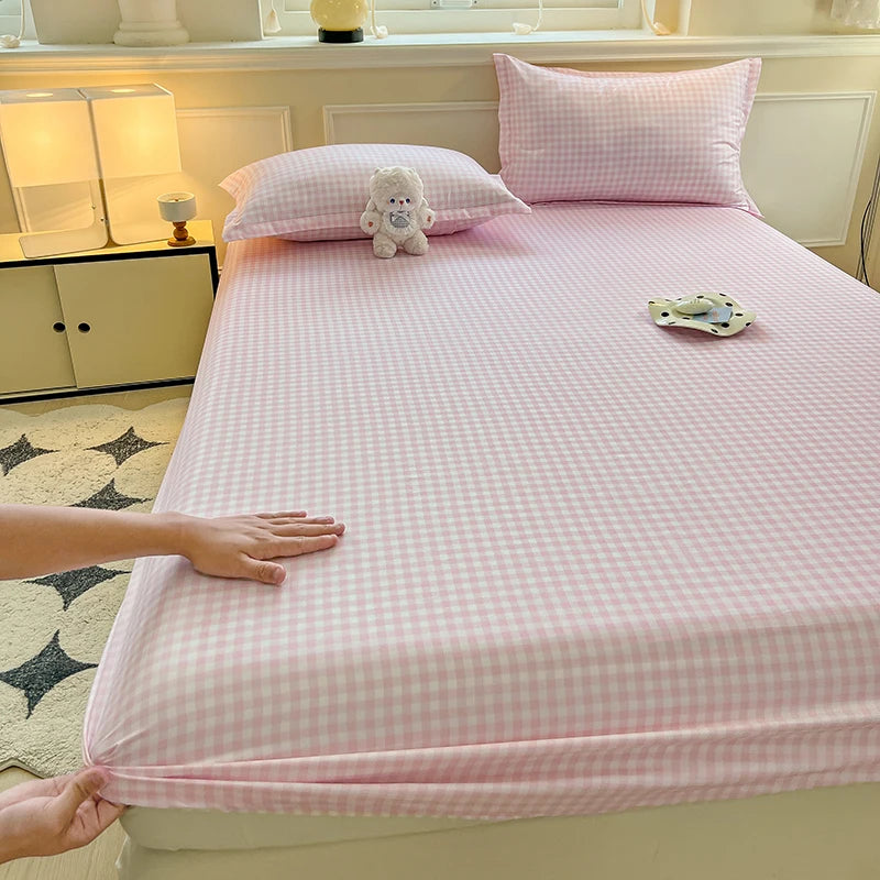 A close-up view of a hand smoothing a soft pink and white plaid fitted sheet onto a mattress. The room features warm lighting and minimalist decor.