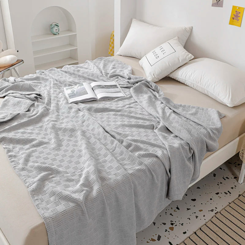 A silver grey textured blanket draped over a bed with white pillows and an open book.