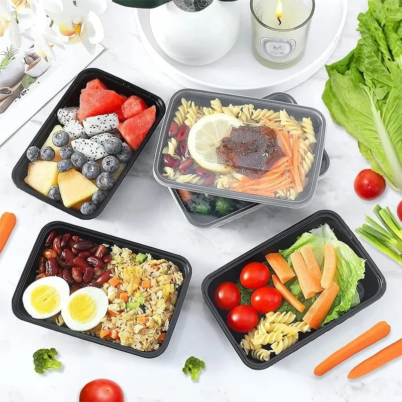 Prepared meals in black containers on a white table with vegetables and fruits.