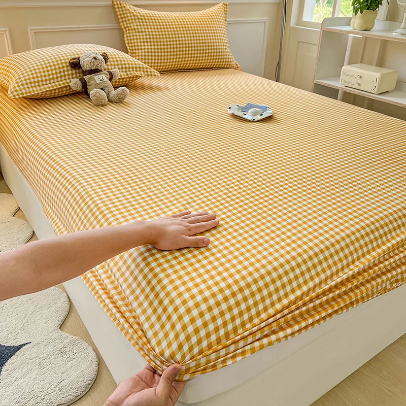 A high-angle shot of a bed featuring a yellow and white mini-plaid gingham fitted sheet and matching pillowcase. A small plush dog toy sits on the pillow against a cream-colored headboard.