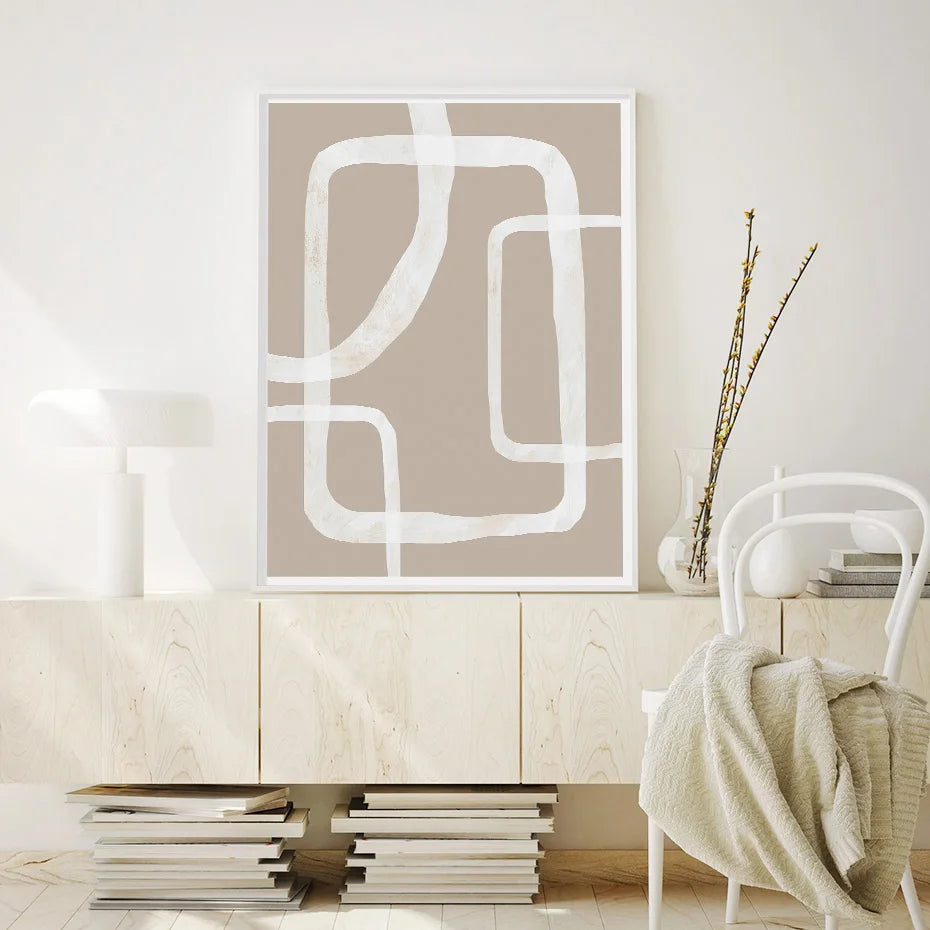 Framed abstract wall art with white curved line shapes on a warm beige background, hanging above a light wood bench in a softly lit interior with a vase of dried grass nearby.