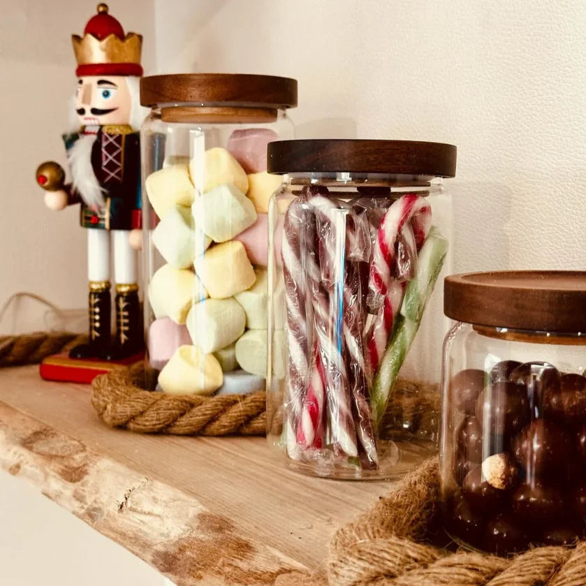 Three glass jars with wooden lids filled with various candies on a wooden surface, with a nutcracker figurine in the background.