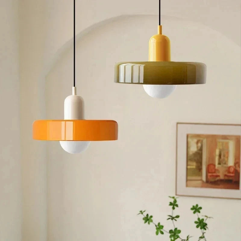 Two modern pendant lights with disc-shaped shades hang in a softly lit room. One light has an orange shade and a white stem, while the other has an olive green shade and a yellow stem.