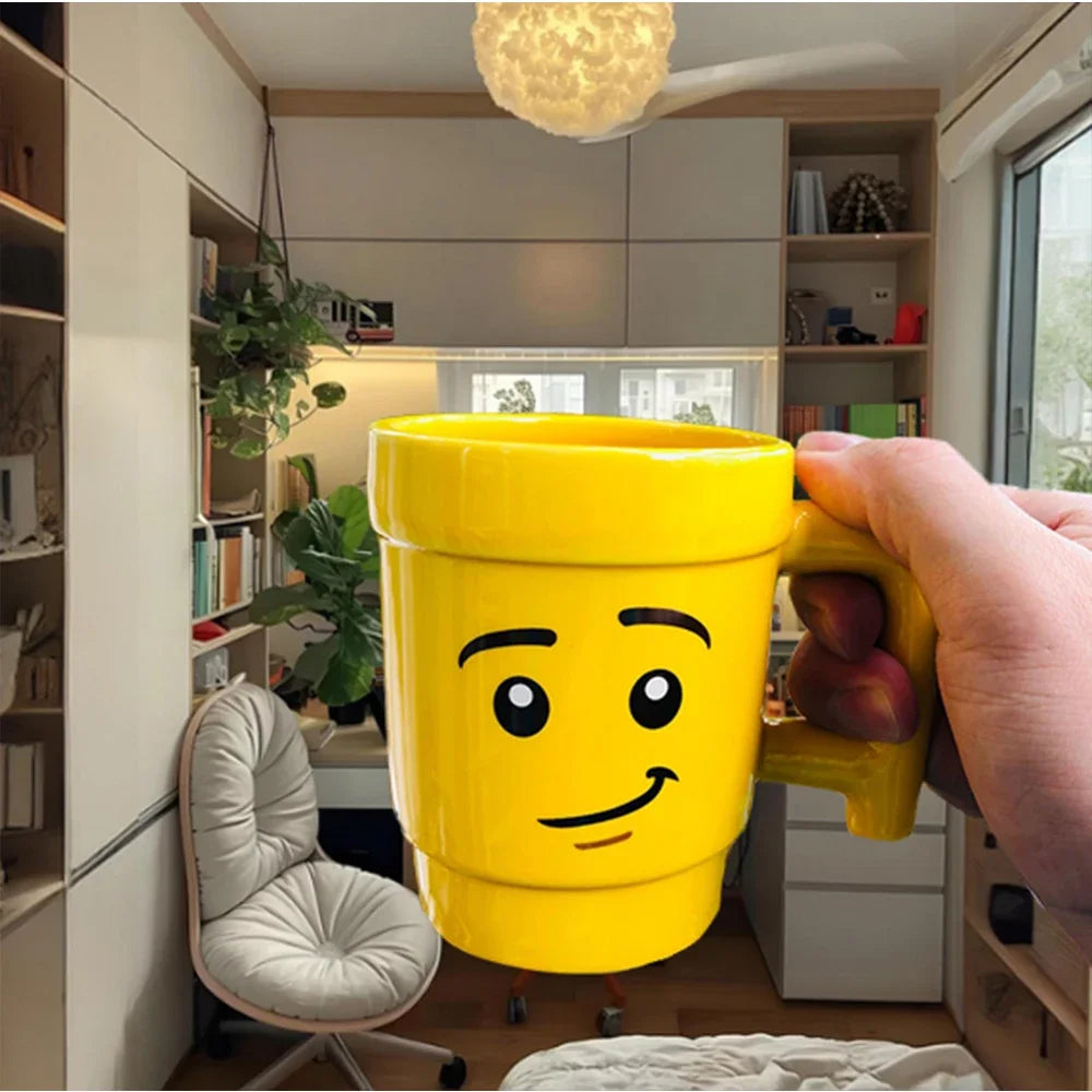 Yellow mug with a face design held in a room with bookshelves and a chair.