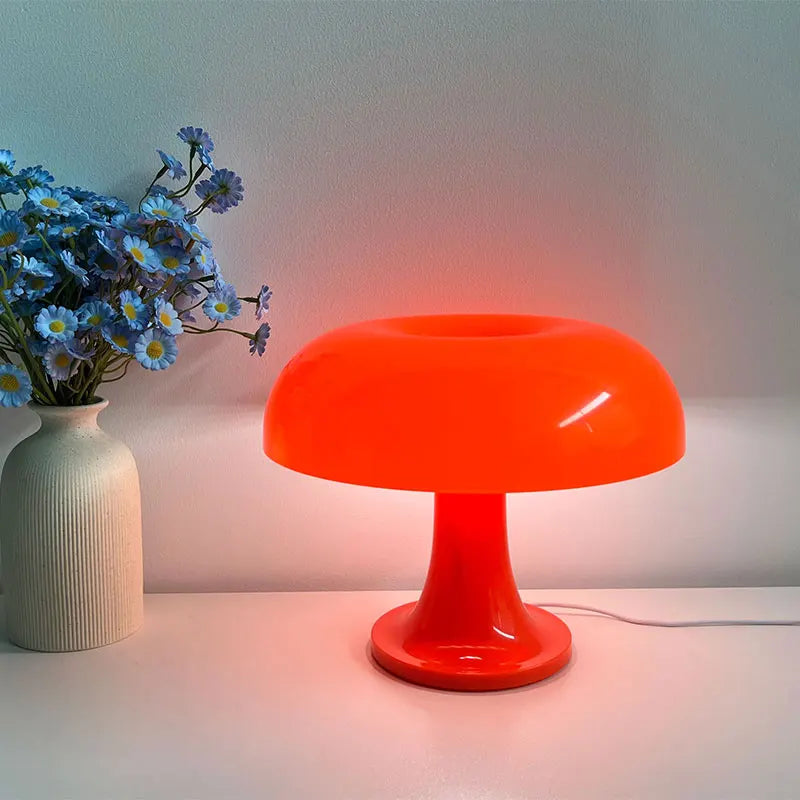 Orange mushroom-shaped table lamp on a white surface with a vase of blue flowers next to it.