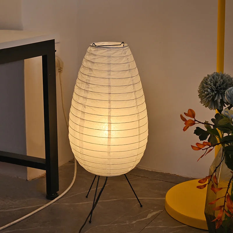 White paper lantern on a stand in a room setting with a plant and table.