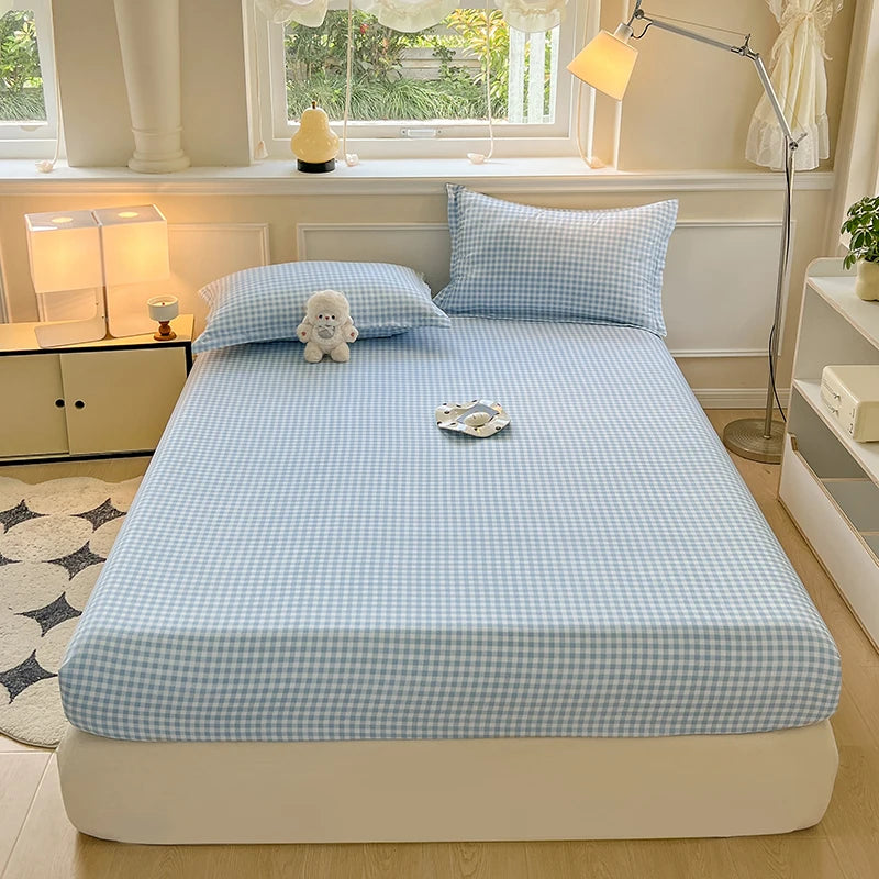 A bed made with a light blue and white gingham check fitted sheet and pillowcase. A small white plush bear is resting on the pillow in a brightly lit, aesthetic room.