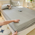 A bed featuring a muted sage green and grey plaid fitted sheet. The fabric has a 300-thread-count texture, shown with a matching pillowcase and neutral room accents.