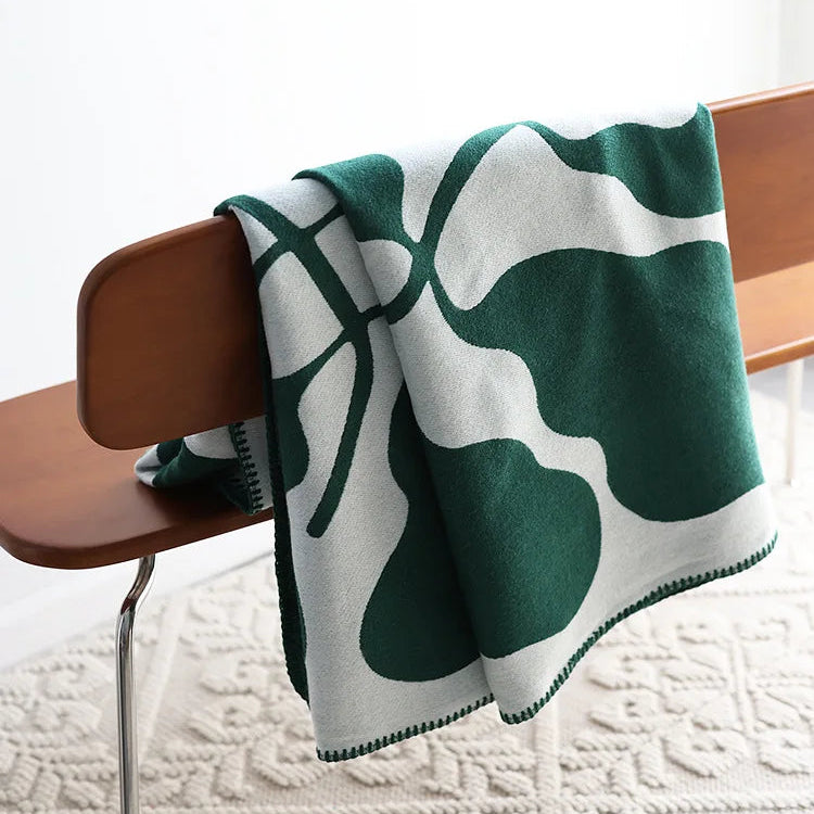 Forest green and white abstract jacquard blanket with blanket stitch edge draped over a wooden chair on a textured cream rug