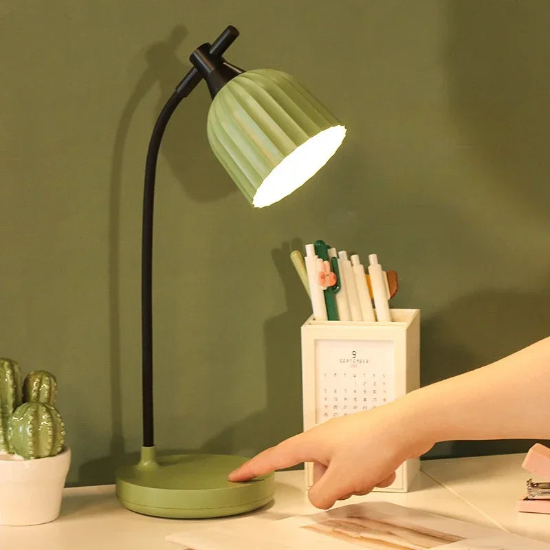 Green desk lamp on a table with a hand reaching towards it against a green wall.