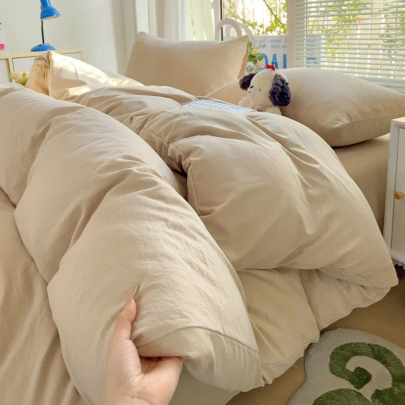 Beige comforter being held over a bed in a bright room with toys on the floor.