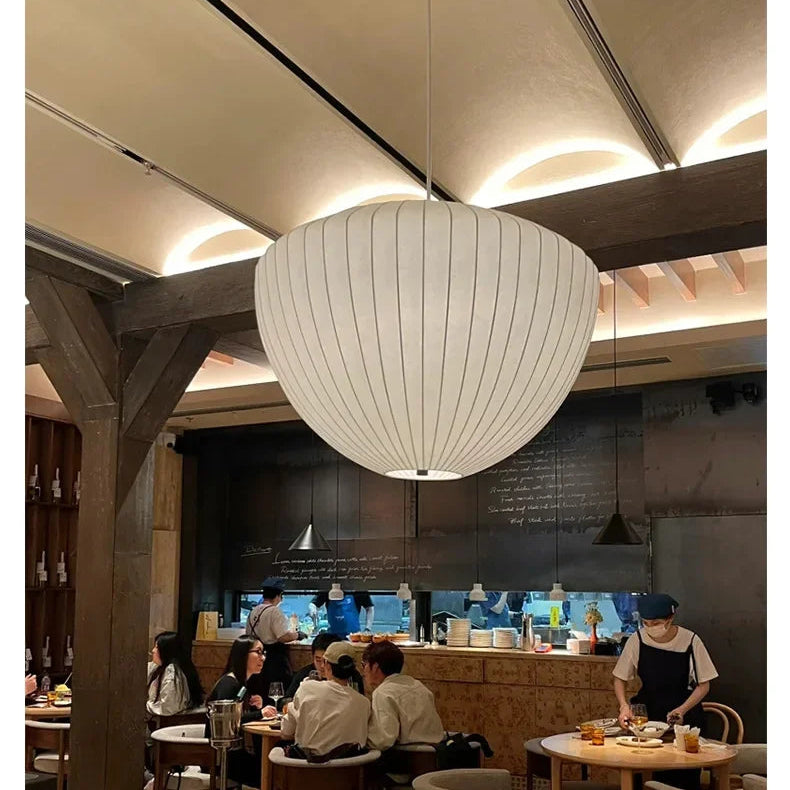 Dining area with a large white pendant light, people seated at tables, and a kitchen in the background.