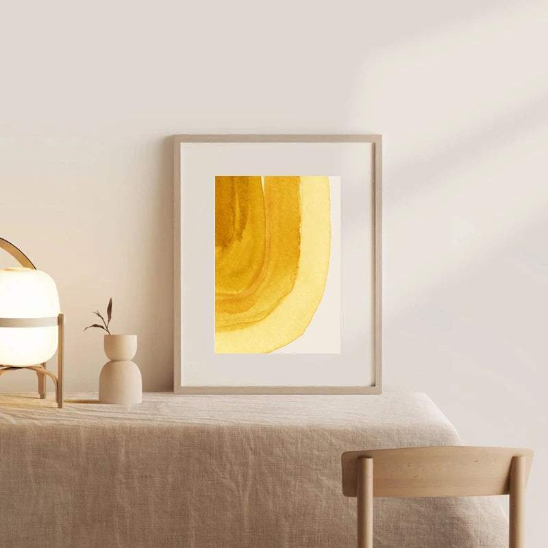 Framed abstract yellow artwork on a wall above a table with a lamp and vase.