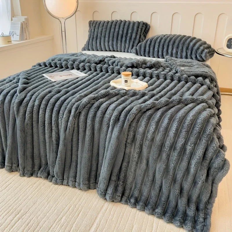 A grey textured blanket draped over a bed with pillows and a small table in a bedroom setting.
