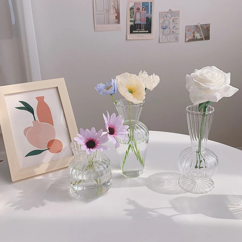 Three clear glass vases with flowers on a white surface, framed artwork in the background.
