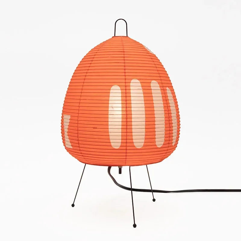 An orange ribbed paper table lamp with a teardrop shape, supported by three thin black metal legs and a black power cord, shown against a plain white background.