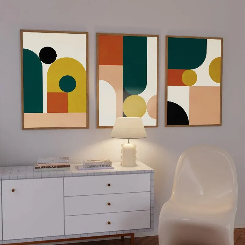 Three abstract art pieces on a wall above a white cabinet with a lamp.