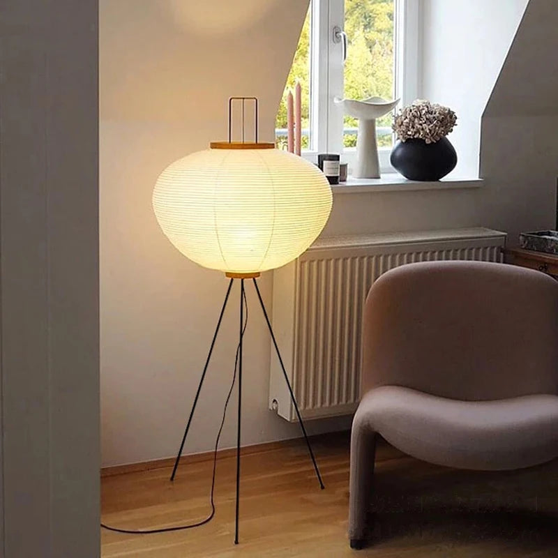 A Japanese floor lamp with a round white shade in a room with a window and a chair.